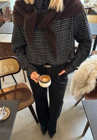 Personne portant une chemise à carreaux et un pantalon noir, tenant une tasse de café avec un art latte, debout dans un café avec des chaises et des tables en bois à proximité.