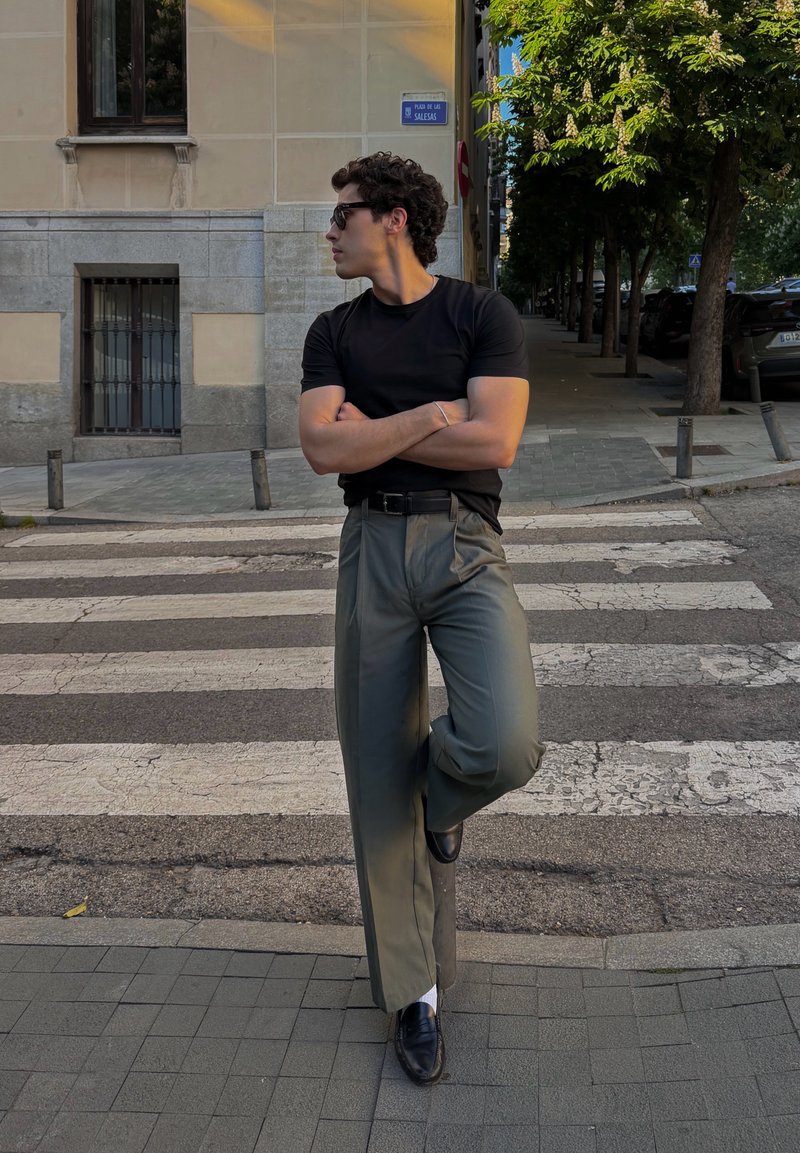 Homme portant des lunettes de soleil, une chemise noire et un pantalon gris, debout les bras croisés et un pied levé dans une rue de la ville près d'un passage pour piétons.