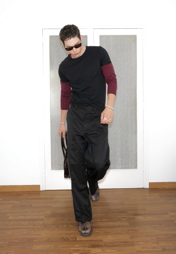 Hombre con gafas de sol, camisa de capas negra y granate, pantalones negros y botas, caminando en interiores sosteniendo una bolsa negra.