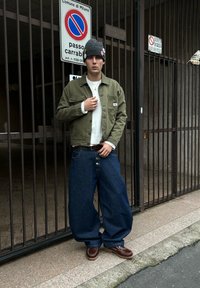 Jeune homme portant un bonnet gris, une veste olive, un pull blanc, un jean bleu surdimensionné et des chaussures marron, debout près d'une entrée grillagée à Milan.