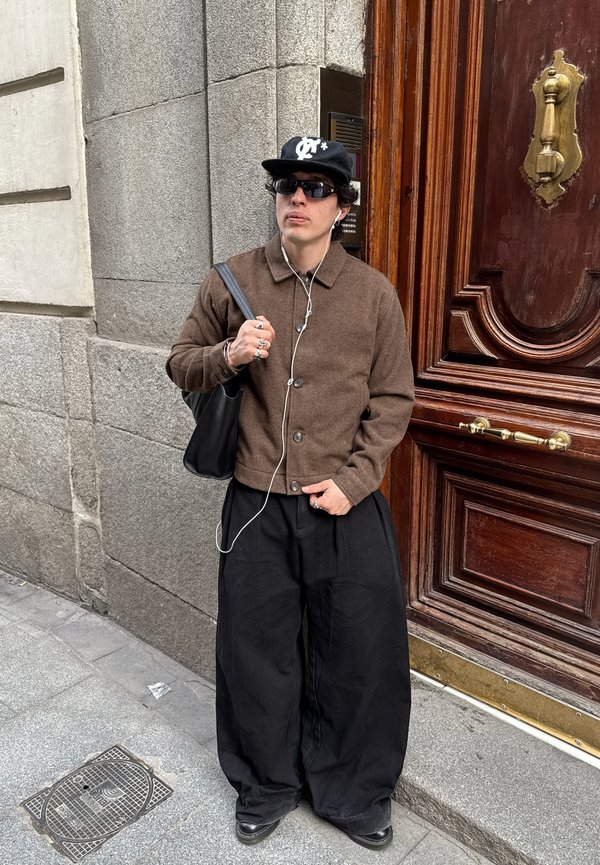 Jeune personne en veste marron, pantalon large noir, casquette noire et lunettes de soleil, debout près d'une grande porte en bois avec des poignées en laiton, tenant un sac noir.