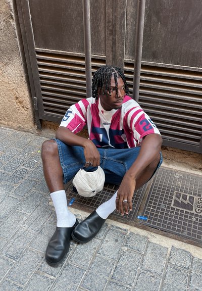 Camisa de manga corta a rayas en rojo, blanco y azul, combinada con pantalones cortos de mezclilla. Calzado negro, calcetines blancos, sosteniendo una gorra de color claro.
