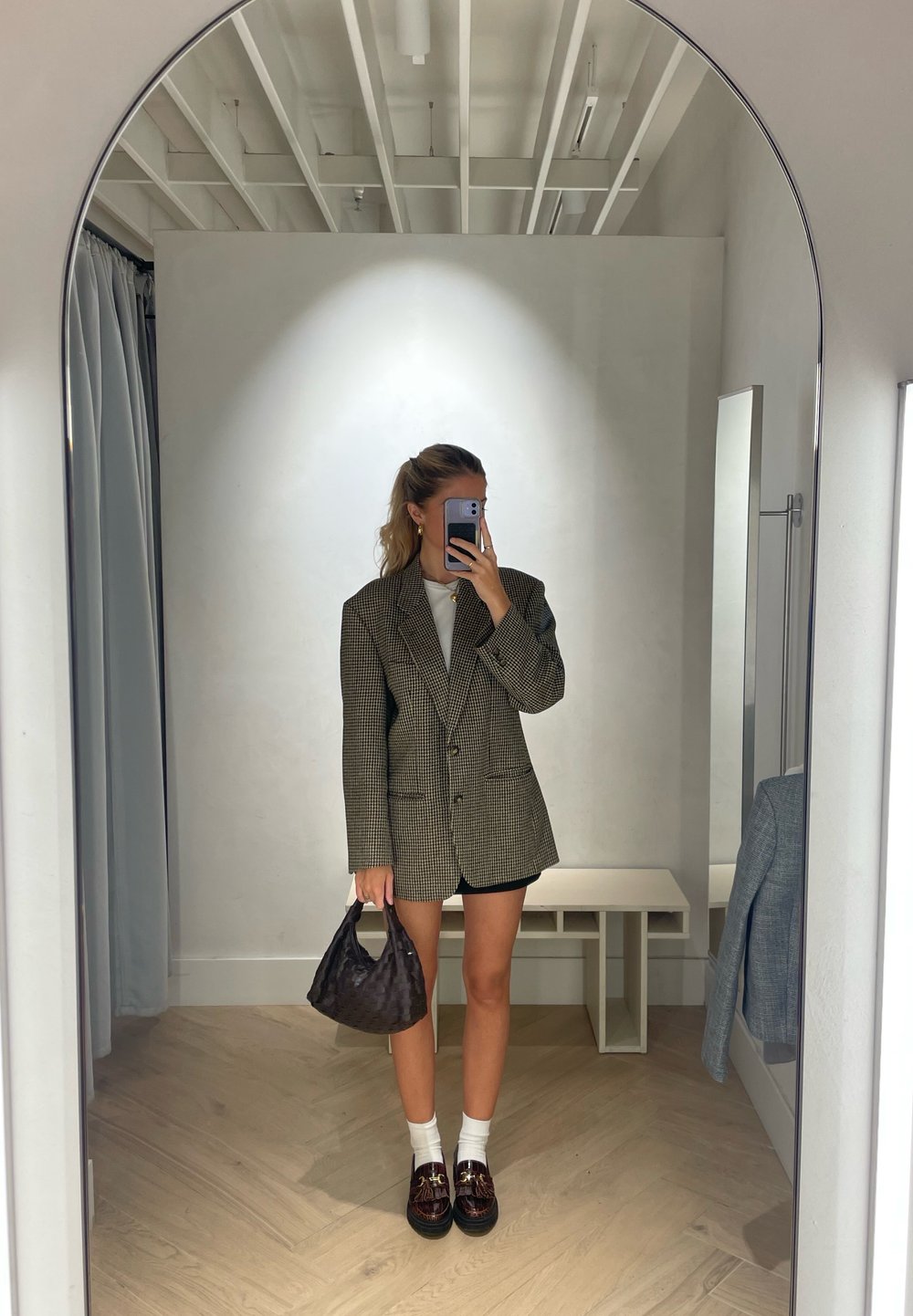Blazer oversized xadrez em cinza e preto, combinado com uma blusa branca, calções pretos e uma bolsa de mão de tecido escuro. Sapatos de plataforma com detalhes.