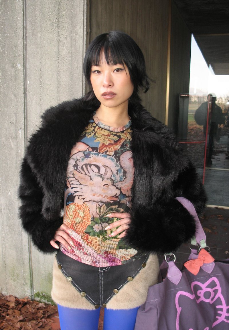 Giovane donna con giacca di pelliccia nera, top con stampa colorata di draghi, gonna bordata di pelliccia, calze blu e una borsa viola di Hello Kitty.