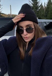 Pessoa a usar um gorro preto e óculos escuros escuros, vestida com uma jaqueta de fleece azul-marinho sobre uma blusa preta, num ambiente ao ar livre com árvores.