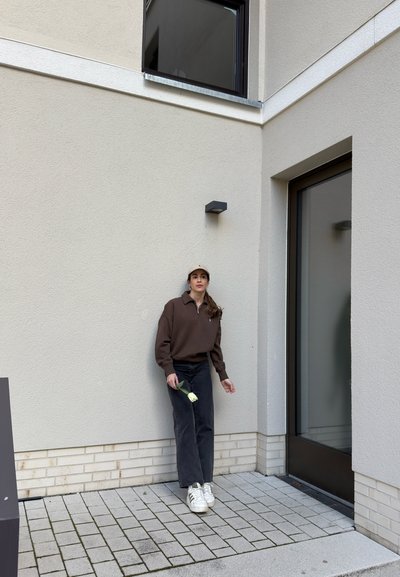 Mujer con sudadera marrón y gorra sosteniendo un tulipán blanco, de pie junto a una pared beige cerca de una puerta y ventana de cristal en un patio exterior.
