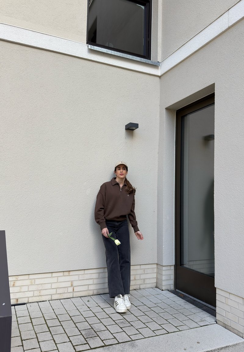 Frau in braunem Sweatshirt und Kappe hält eine weiße Tulpe und steht vor einer beigen Wand nahe einer Glastür und einem Fenster in einem Außenhof.