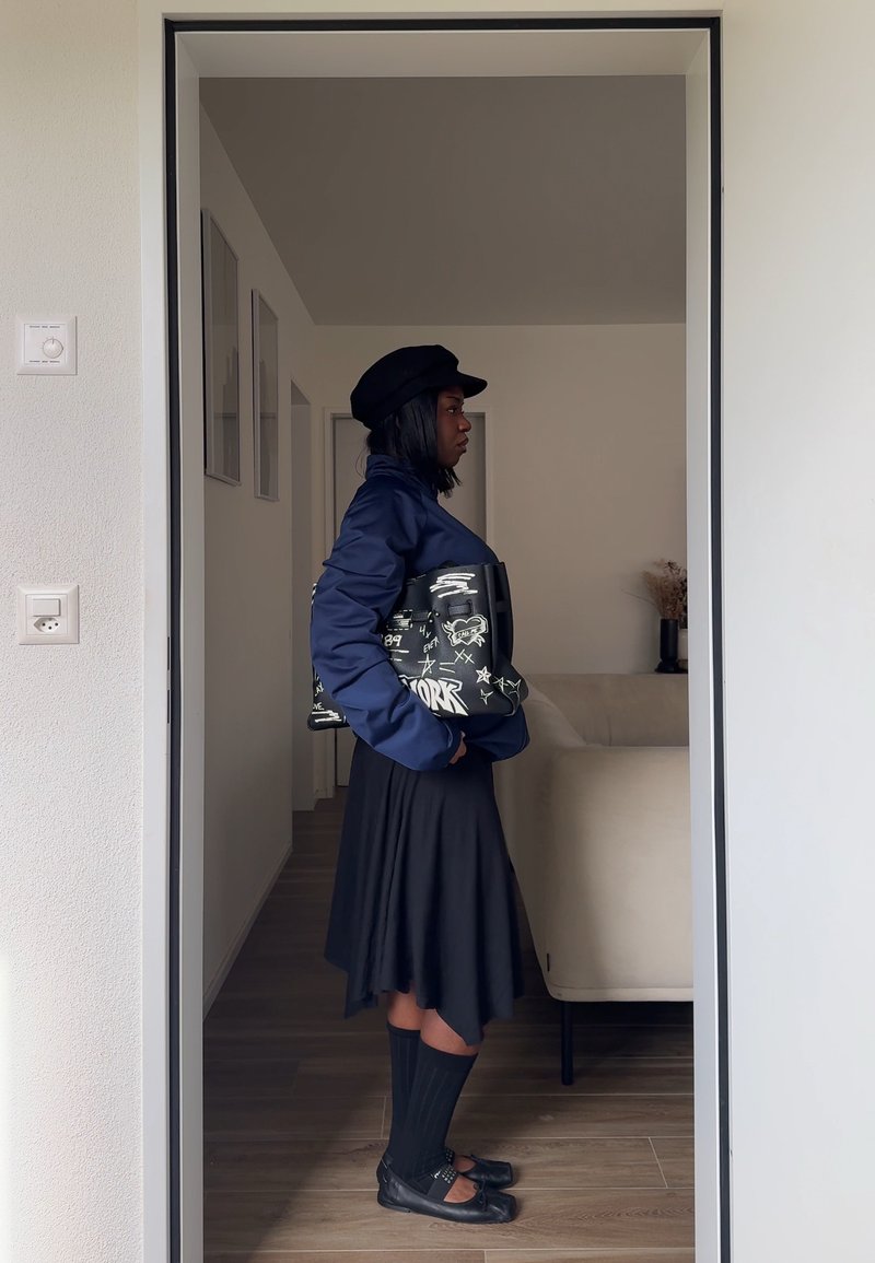 Femme en veste bleu marine, jupe noire, chaussettes hautes et chapeau, tenant un sac noir à motifs, debout dans l'embrasure d'une porte à l'intérieur d'une maison moderne.