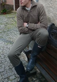 Jeune personne assise sur un banc portant un pull marron, un pantalon gris, des bottes de pluie Hunter noires et une montre-bracelet, en plein air sur un chemin pavé.