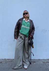 Πράσινη φούτερ μπλούζα Adidas πάνω από ένα λευκό πουκάμισο με γιακά, καφέ δερμάτινο μπουφάν, μπεζ παντελόνι σε φαρδύ σχέδιο, μαύρη τσάντα και λευκά αθλητικά παπούτσια.