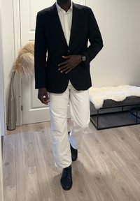 Schwarze Blazer über einem weißen Hemd, kombiniert mit weißen Hosen und schwarzen Schuhen. Glatter Stoff und figurbetontes Design auf hellem Holzboden.