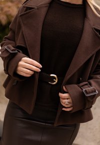 Ruskea villasekoitteinen takki leveillä kauluksilla, kireillä hihoilla ja mustalla nahkavyöllä, jossa on kultainen solki, tummanruskean villapuseron ja nahkahameen päällä.