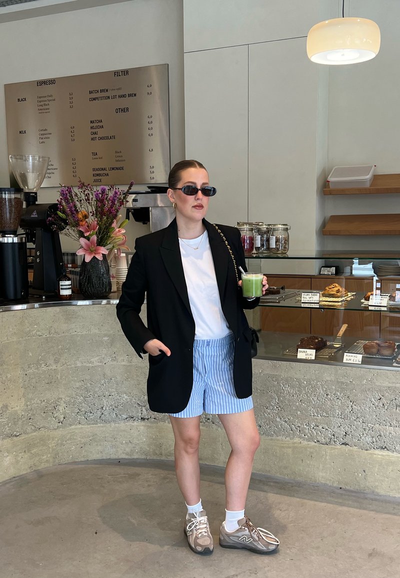 Veste noire oversize, t-shirt blanc, short rayé bleu, et baskets beiges. Tenant une boisson verte, debout dans un café avec des pâtisseries.