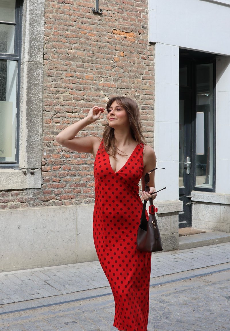Femme en robe rouge à pois tenant des lunettes de soleil et un sac brun, marchant sur une rue pavée à côté de bâtiments en brique et en pierre.