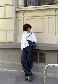 Persoon met krullend donker haar in een witte lange mouwen shirt, brede donkere jeans en versierde zwarte schoenen, staand tegen een beige muur van een gebouw.