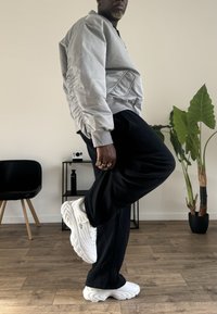 Veste bomber gris clair avec manches froncées, pantalon foncé et baskets blanches sculpturales. Cadre intérieur avec une plante et du mobilier.