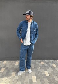 Jeansjacke und Jeanshose in Blau, übereinander getragen mit einem weißen T-Shirt. Enthält eine graue Mütze mit Logo und weiße Turnschuhe. Steht auf gemustertem Pflaster.