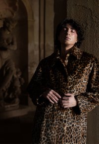 Personne aux yeux fermés portant un manteau à imprimé léopard, appuyée contre un mur texturé dans un intérieur orné faiblement éclairé avec une statue en pierre à proximité.