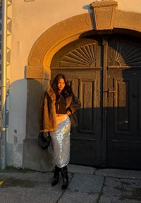 Veste en fausse fourrure marron, crop top noir, jupe midi à sequins, bottes noires montantes jusqu'aux genoux et petit sac à main noir, posés devant des portes en bois.