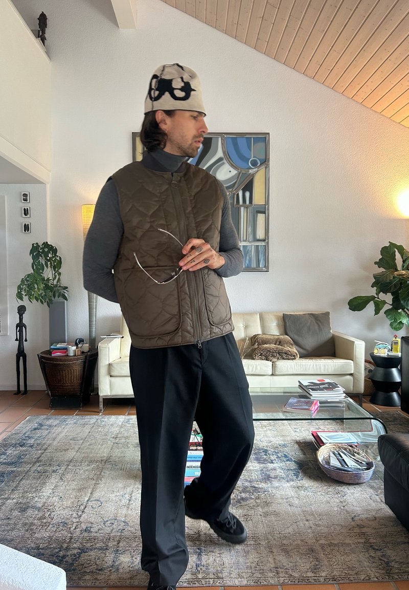 Veste matelassée marron portée sur un col roulé foncé, pantalon noir, tenant des lunettes ; chapeau blanc avec motifs noirs ; cadre intérieur avec des plantes.