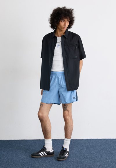 Camisa negra de botones, camiseta blanca, pantalones cortos azul claro con rayas negras, deportivas negras con rayas blancas, calcetines blancos de caña corta y tatuajes visibles.