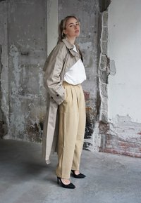 Casaco trench bege sobre uma camisa branca, combinado com calças bege de cintura alta e saltos agudos pretos. Paredes cinzentas texturizadas ao fundo.
