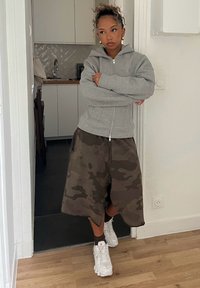 Šedá mikina na zip, maskáčové šortky a bílé tenisky s akcenty. Pozadí tvoří světlé stěny a kuchyňské skříňky.