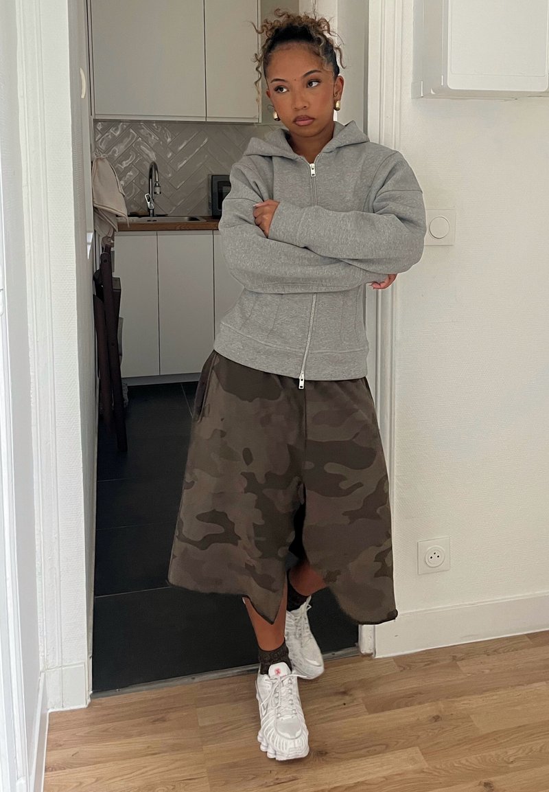 Haut à capuche gris zippé, shorts en camouflage et baskets blanches avec des accents. L'arrière-plan présente des murs clairs et des meubles de cuisine.