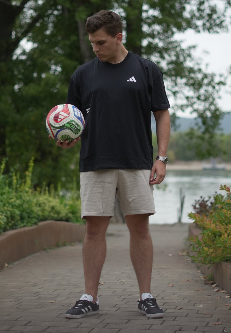Jeune homme en chemise noire et short beige tenant un ballon de football en plein air sur un chemin pavé près de l'eau et de la verdure.