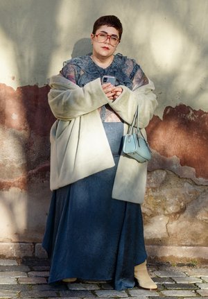 Persona con cabello corto y gafas, vistiendo una blusa azul de encaje, falda larga azul, abrigo beige y sosteniendo un teléfono y un bolso pequeño al aire libre.
