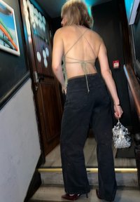 Jean noir à jambes larges associé à un haut vert clair à bretelles. Un sac transparent avec des accents argentés est tenu dans une main. Fond en bois et mur.