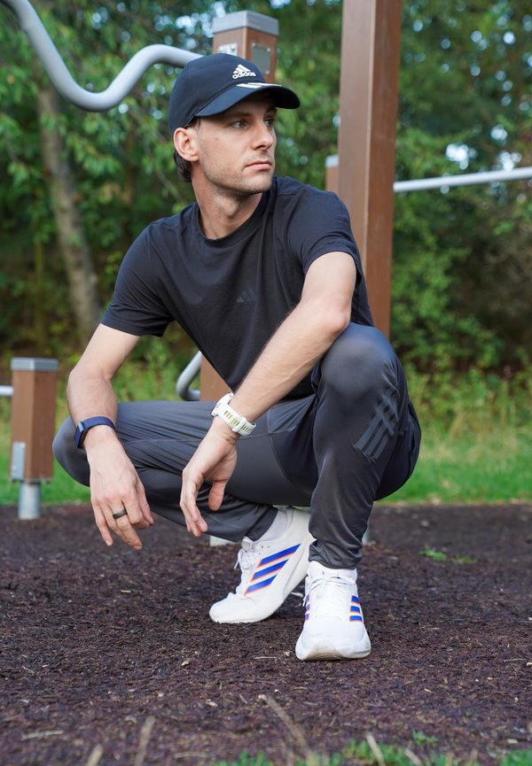 Homme en tenue de sport Adidas noire et casquette fait des squats sur du paillis sombre dans un parc de fitness en plein air avec de la verdure en arrière-plan.