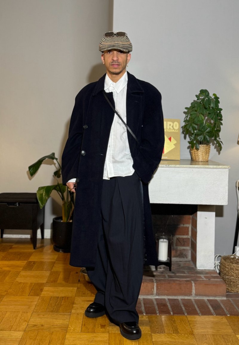 Homme portant un pardessus noir, une chemise blanche, un pantalon large noir, des chaussures noires et une casquette à carreaux, debout à l'intérieur près d'une cheminée et de plantes en pot.