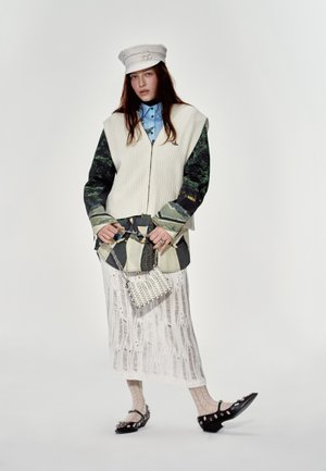 Gilet crème texturé sur des manches à motifs, chemise bleue, jupe blanche en dentelle, collants en résille à motifs, chaussures noires et un petit sac en perles.