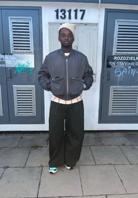 Sivá bomber bunda so zipsovými vreckami, pruhované tričko, široké zelené nohavice a viacfarebné tenisky na dlažobných kockách. Mestské pozadie.