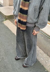Pantalon en denim gris délavé à jambes larges avec ourlets effilochés, associé à un pull gris texturé et une écharpe rayée multicolore. Chaussures marron.