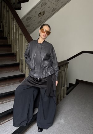 Mujer con chaqueta de cuero oscura de gran tamaño, pantalones anchos negros y grandes gafas de sol color ámbar, de pie en unas escaleras de madera en el interior.