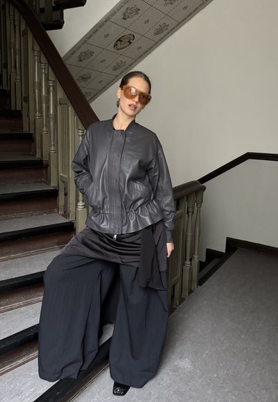 Mujer con chaqueta de cuero oscura de gran tamaño, pantalones anchos negros y grandes gafas de sol color ámbar, de pie en unas escaleras de madera en el interior.