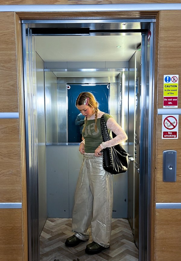 Noor naine punutud juustega seisab lifti sees, kohendab pükse, kannab musta õlakotti, väljas on hoiatus- ja suitsetamine keelatud märgid.