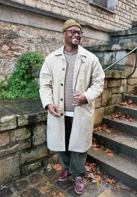 Beige trench coat, striped jumper, green corduroy trousers, brown boots; standing on stone steps with autumn leaves, natural background.