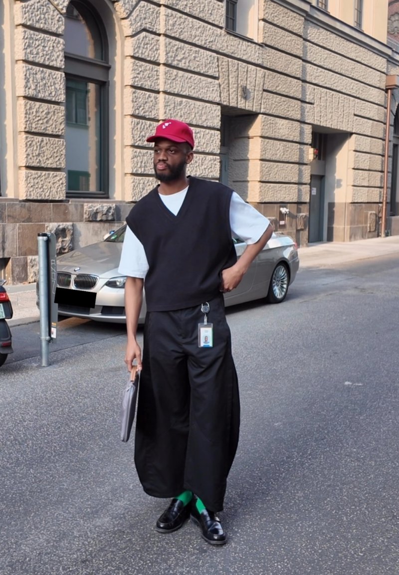 Pull noir sans manches sur une chemise blanche, pantalon noir à jambes larges, chaussettes vertes et chaussures noires, tenant un objet argenté. Casquette rouge et badge d'identification visibles.