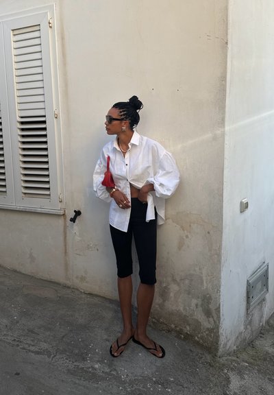 Camisa blanca de gran tamaño con botones y mangas abullonadas, combinada con leggings capri negros y chanclas negras, sosteniendo un pequeño bolso rojo.