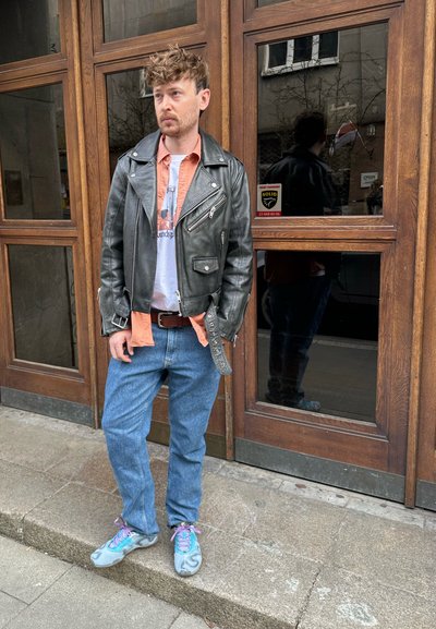 Chaqueta de moto de cuero negra sobre una camiseta naranja, camiseta gráfica blanca, jeans azules y zapatillas grises con cordones morados. Fondo de paneles de madera.