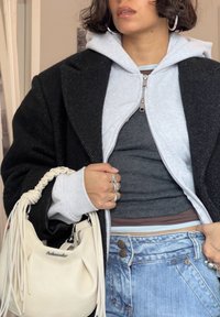Manteau à chevrons noir sur un sweat à capuche zippé gris clair, débardeur gris superposé, jean déchiré taille haute, sac à main blanc avec détails à franges.