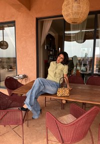 Ein hellgrünes Oberteil mit einer Schleifendetail, hellblaue Jeans mit weitem Bein und eine schwarze Handtasche mit einem goldgemusterten Design, auf einem Holztisch im Freien arrangiert.