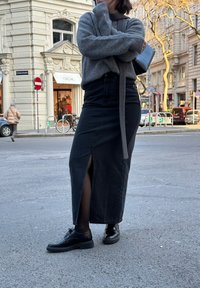 Šedý pletený svetr, černá džínsová sukně s bočním rozparkem, černé kožené boty a malá černá taška. Pozadí zahrnuje městskou ulici.