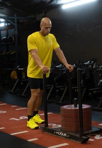 Rumena športna majica, črne kratke hlače in neonsko rumeni športni copati. Potiska sani z naloženimi utežmi po telovadniški podlagi.