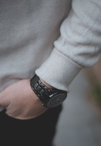 Μαύρο μεταλλικό ρολόι με στρογγυλή πρόσοψη και ασημί λεπτομέρειες, φορεμένο πάνω από έναν ανοιχτό γκρι πλεκτό πουλόβερ. Το χέρι resting σε μια τσέπη.