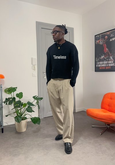 Hombre con gafas y pelo trenzado que lleva un suéter negro "Timeless" y pantalones beige, de pie en una habitación con una planta, una silla naranja y un póster de baloncesto.