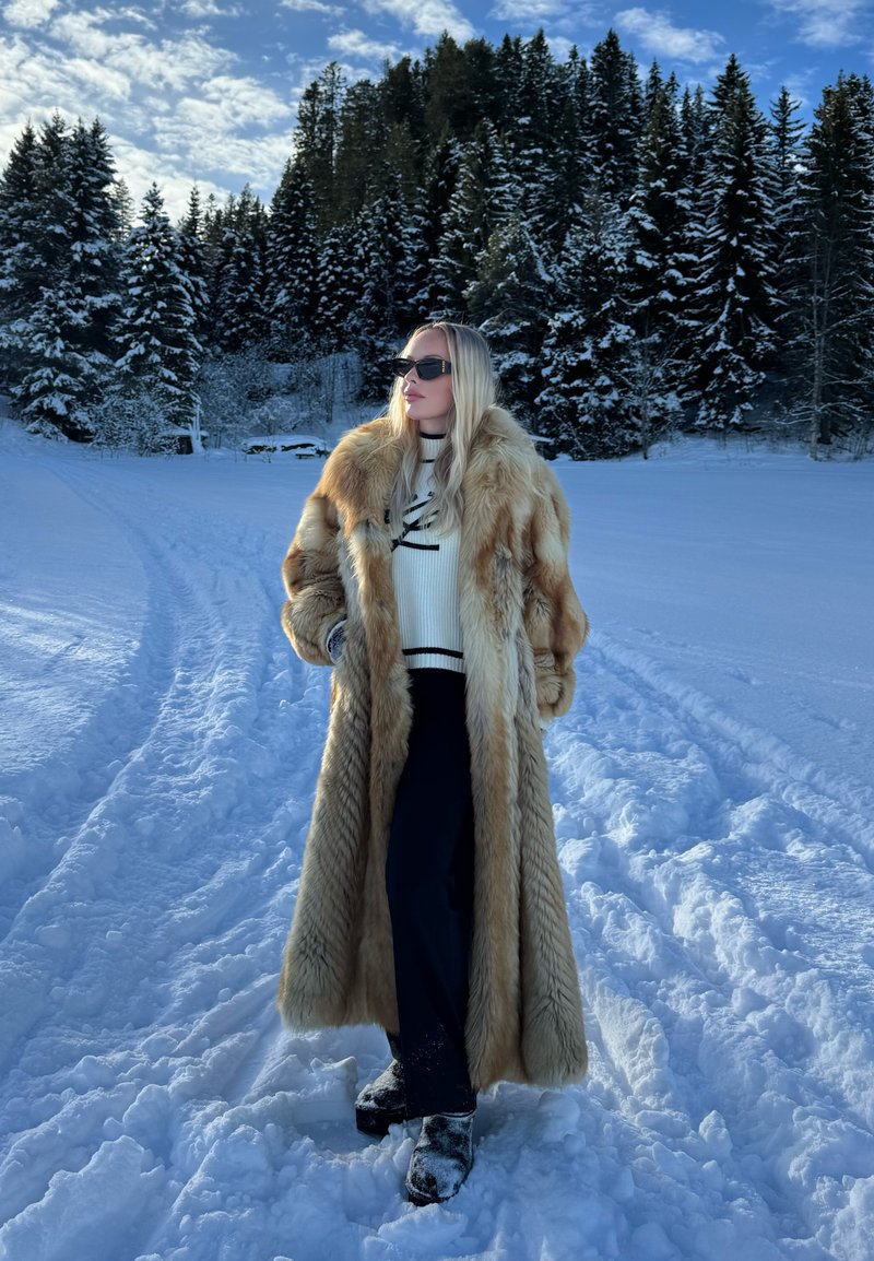 Femme portant un long manteau en fourrure beige, pantalon noir, lunettes de soleil, debout sur un sol enneigé avec une forêt en arrière-plan sous un ciel bleu.